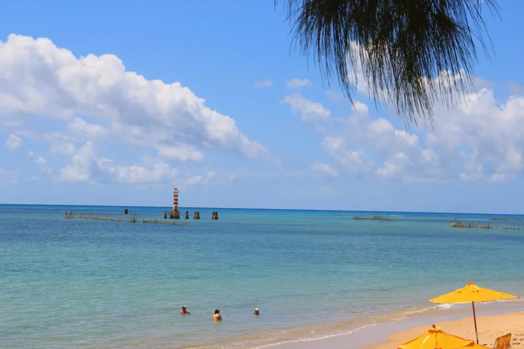 Praia de Pajuçara em Maceió com farol e mar cristalino sob guarda-sol amarelo